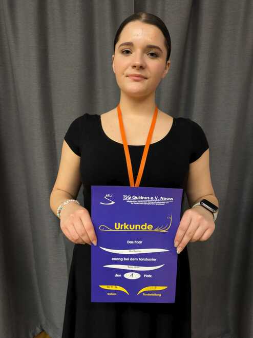 Mädchen mit orangefarbener Medaille hält eine Urkunde für den ersten Platz beim Tanzturnier vor grauem Hintergrund.