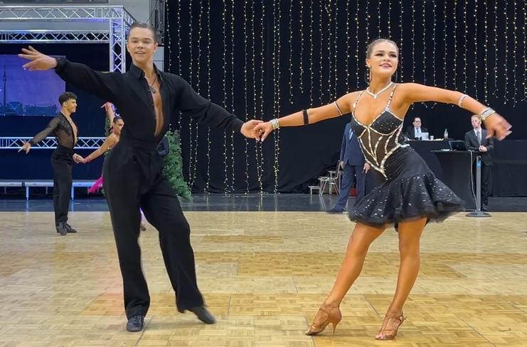 Tanzpaar in eleganter Kleidung vollzieht einen Schritt auf einer Tanzfläche mit festlicher Beleuchtung im Hintergrund.