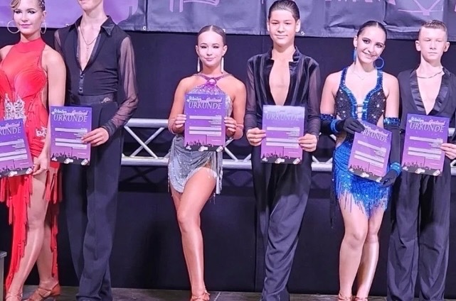 Fünf Tänzer in eleganter Kleidung halten Urkunden bei einem Wettbewerb. Dahinter ist eine Bühnenkulisse sichtbar.