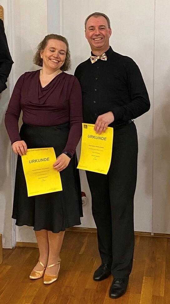 Paar in formeller Kleidung hält Urkunden in der Hand und steht lächelnd vor einer weißen Wand.