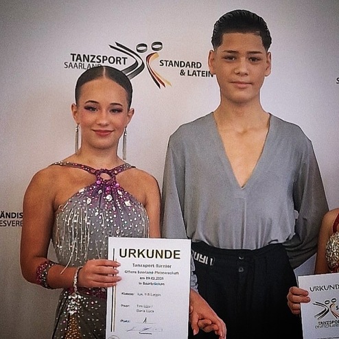Zwei Tänzer stehen mit einer Urkunde in der Hand vor einem Hintergrund mit Tanzsport-Logo.