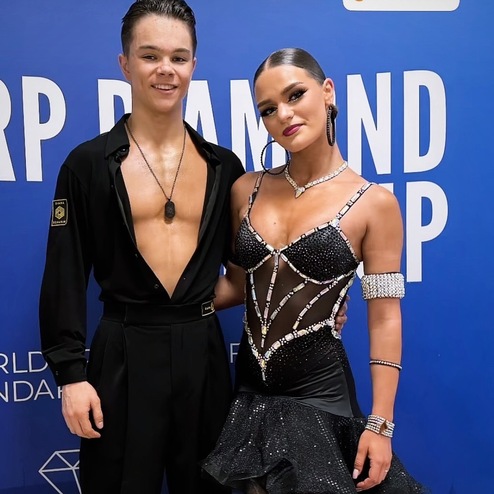 Paar in eleganter Tanzkleidung steht vor einem blauen Hintergrund mit Aufschrift „Weltmeisterschaft“.