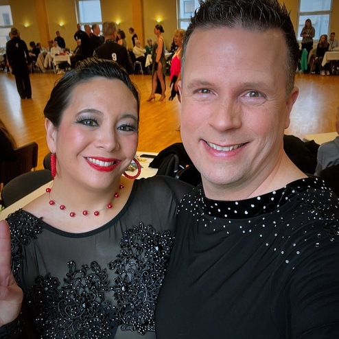 Selbstporträt eines lächelnden Paares in festlicher Tanzkleidung, vor einem hellen Veranstaltungsort mit Tänzern im Hintergrund.