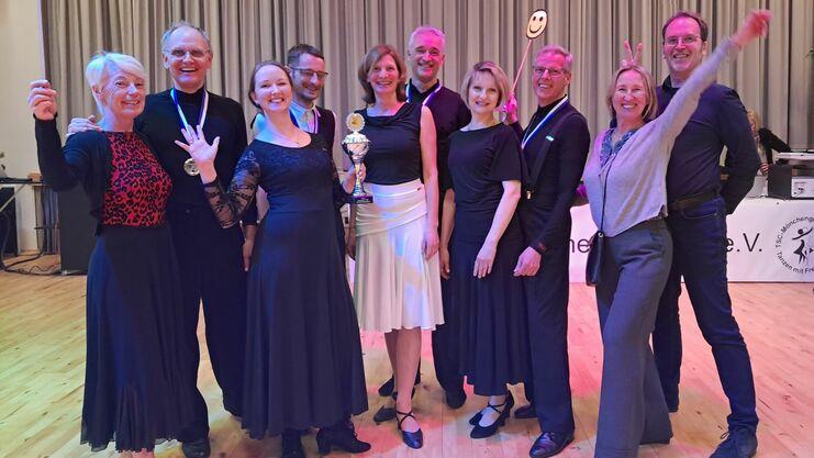Gruppe von Tänzern in eleganter Kleidung, die mit Trophäe und lächelnden Gesichtern vor einer Bühne posieren.