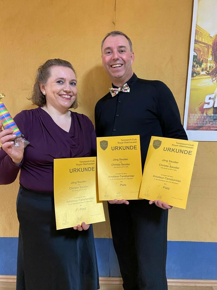 Paar hält drei goldene Urkunden eines Amateur-Tanzturniers vor einer gelben Wand. Frau hält zudem einen bunten Stift.
