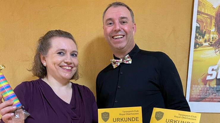 Paar hält drei goldene Urkunden eines Amateur-Tanzturniers vor einer gelben Wand. Frau hält zudem einen bunten Stift.