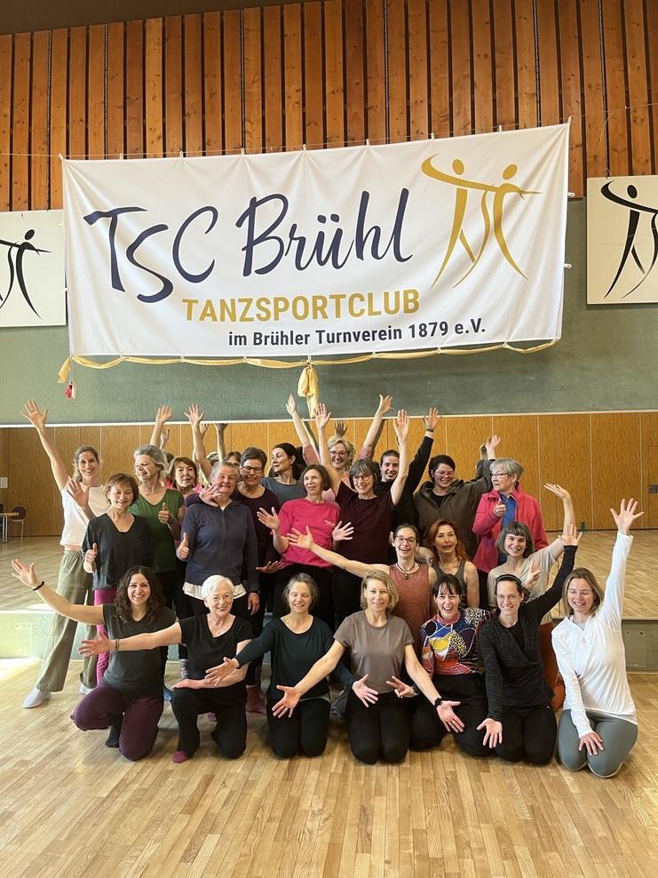 Gruppenkfoto von Mitgliedern des TSC Brühl, die fröhlich posieren und ihre Hände in die Luft heben, in einem Tanzsaal.