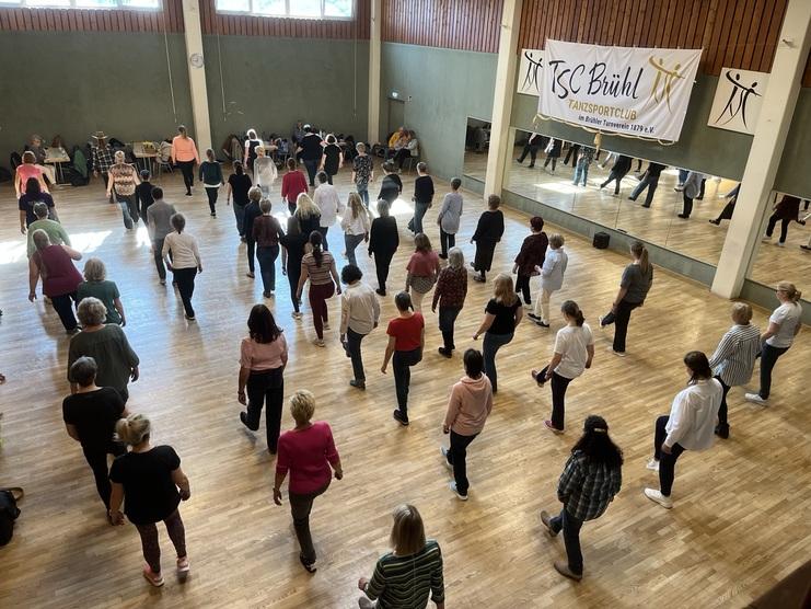 Tanzende Teilnehmerinnen in einem großen Saal, Spiegel und Vereinsbanner im Hintergrund.
