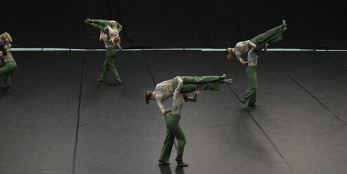 Sechs Tänzerinnen in grüner Kleidung führen akrobatische Lifts auf einer dunklen Tanzfläche aus.