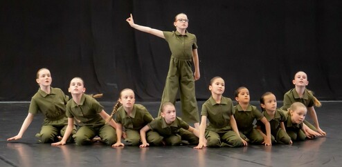 Zehn tanzende Kinder in grünen Overalls, eine hebt den Arm, vor schwarzem Hintergrund.