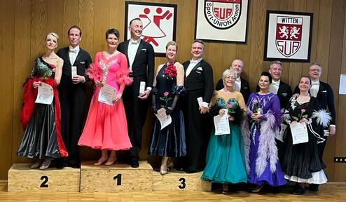 Sieger des Tanzwettbewerbs stehen auf Podesten, tragen elegante Kleider und halten Urkunden in der Hand.