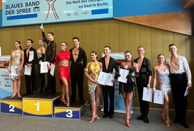 Sieben Tänzerpaare stehen stolz auf dem Siegertreppchen mit Urkunden bei einem Tanzturnier in Berlin.