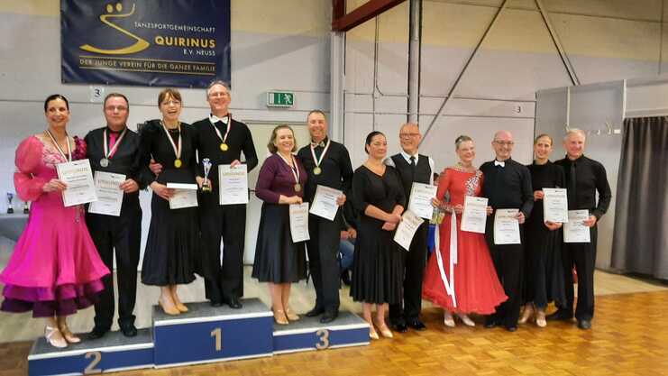 Sieben Tänzer stehen auf einer Siegerehrung, halten Urkunden und Pokale, in festlicher Kleidung.
