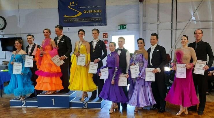 Tanzpaare auf einem Siegertreppchen zeigen Pokale und Urkunden in festlichen Outfits.