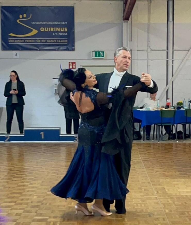 Tanzpaar in eleganter Abendkleidung tanzt im Wettbewerb auf einer Holzfläche, im Hintergrund festliche Tische.