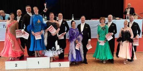 Tanzpaare in festlicher Garderobe stehen auf einer Siegerehrungsbühne, mit Urkunden in der Hand.