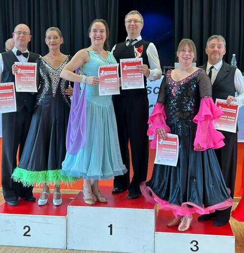Sechs Tänzer in eleganter Wettbewerbsbekleidung halten Urkunden auf einem Siegerpodest mit den Plätzen 1 bis 3.