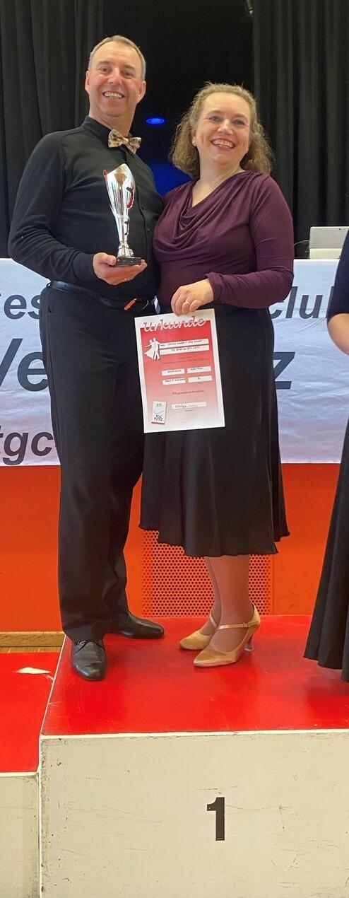 Tänzendes Paar auf dem Siegertreppchen, hält einen Pokal und eine Urkunde, lächelnd in festlicher Kleidung.