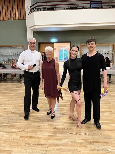 Vier Personen posieren in einem Tanzsaal, zwei Halten Tanzpreise, während hinter ihnen andere Teilnehmer sitzen.