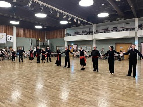 Tanzgruppe in schwarzer Kleidung mit roten Schärpen, die in einem großen Saal eine Tanzaufführung präsentiert.