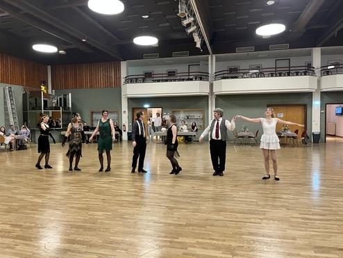 Gruppe von Tänzern in festlicher Kleidung, die auf einer Holzfläche eine Darbietung präsentieren.