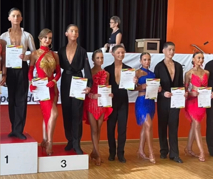 Sechs Tänzer in farbenfrohen Kostümen halten Urkunden bei einem Wettbewerb auf einer Bühne.