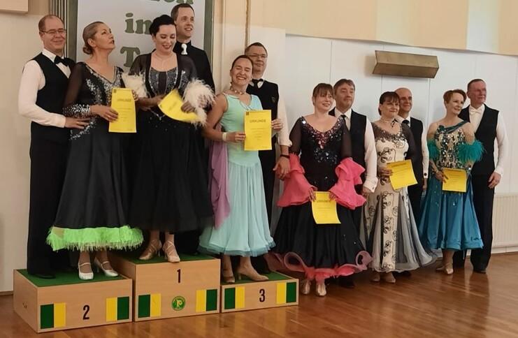 Tanzpaare stehen auf Siegertreppchen, halten Urkunden und tragen festliche Ballkleider und Anzüge in einer Tanzveranstaltung.