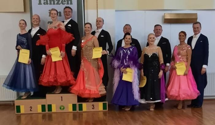 Gruppenfoto von Tänzern in eleganten Kleidern und Anzügen, die auf einem Siegerpodest mit Urkunden posieren.