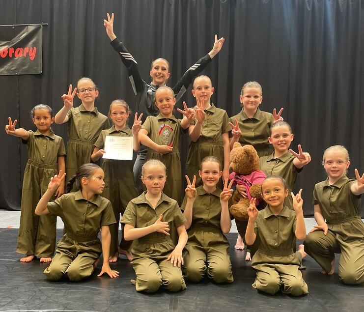 Gruppe von Tänzerinnen in grünen Anzügen posiert lächelnd mit Pokal und Plakat auf einer Bühne.