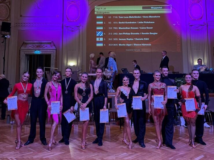 Sechs Paare in farbenfrohen Tanzkostümen halten Urkunden und Medaillen bei einem Tanzwettbewerb in einem festlichen Saal.