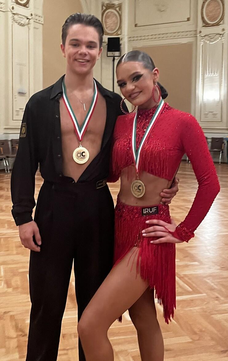 Tanzpaar in Abendkleidung mit Medaillen, stehen in einem festlichen Saal mit Holzboden und elegantem Dekor.