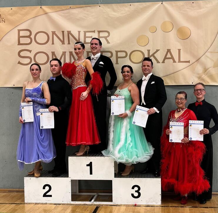 Sieben Tänzer in festlicher Kleidung stehen auf einem Siegertreppchen mit Urkunden vor einem Banner des Bonner Sommertrohpokal.