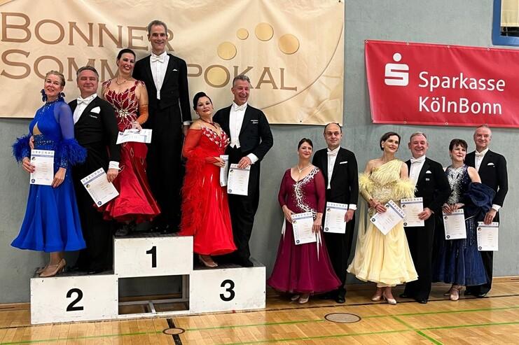 Sieben Paare in eleganter Tanzkleidung stehen auf einem Siegertreppchen bei einem Wettbewerb, Urkunden in der Hand.