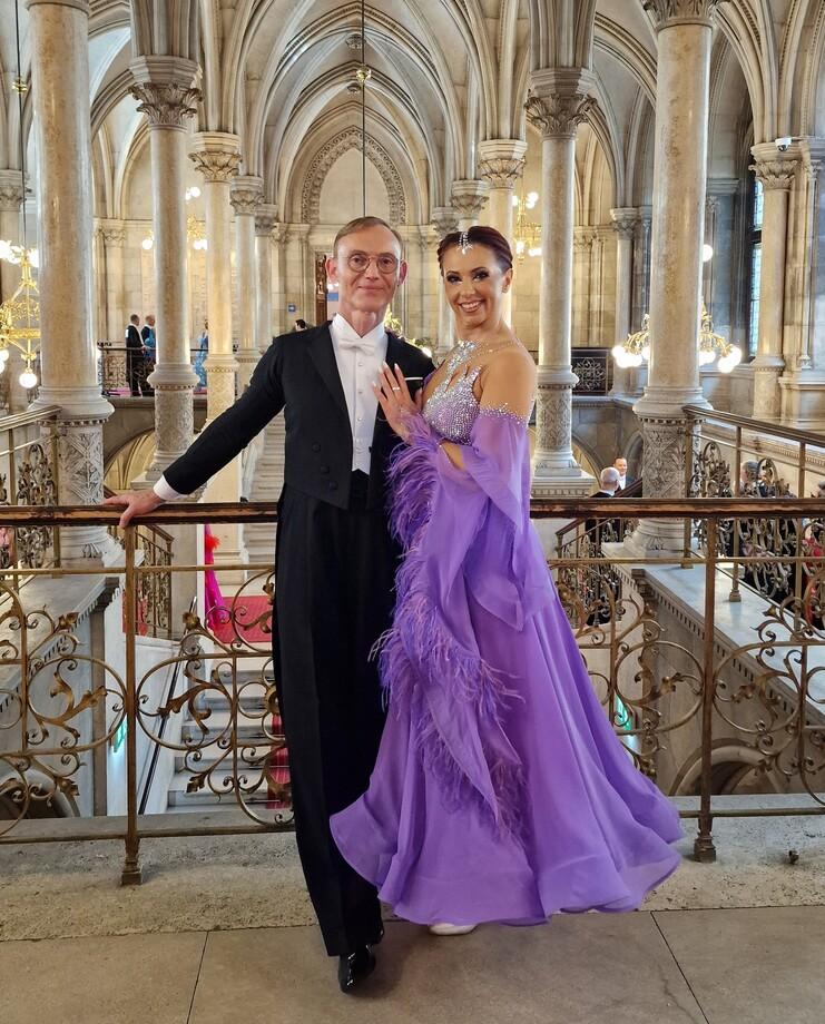 Tanzpaar in eleganter Abendkleidung posiert an einem Balkon mit kunstvoller Balustrade in einem festlichen Saal.