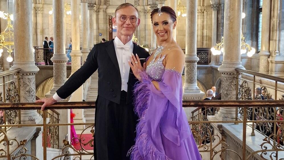 Tanzpaar in eleganter Abendkleidung posiert an einem Balkon mit kunstvoller Balustrade in einem festlichen Saal.