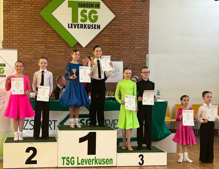 Sechs Kinder in Tanzkleidung stehen auf dem Siegertreppchen und halten Urkunden und Pokale beim Wettbewerb in Leverkusen.