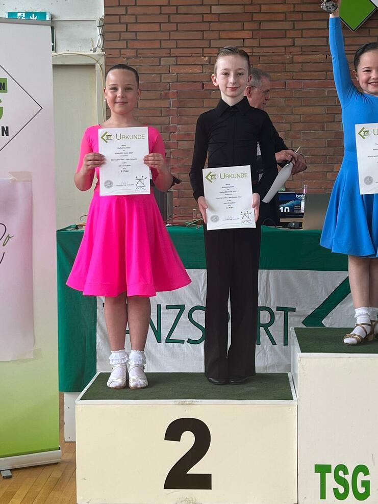 Drei Tänzer stehen auf dem Siegertreppchen, halten Urkunden und tragen bunte Kostüme. Der zweite Platz ist markiert.