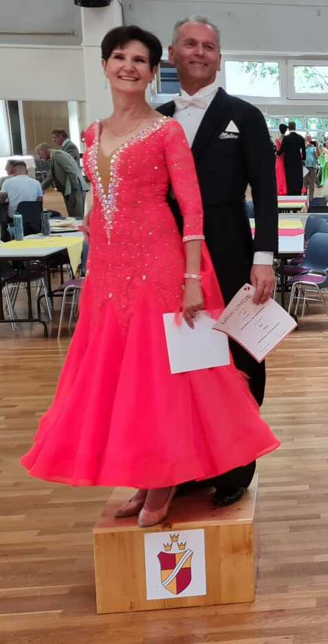 Tanzpaare auf Podesten bei einem Wettbewerb, elegante Kleider, Urkunden in der Hand, festliche Atmosphäre im Hintergrund.