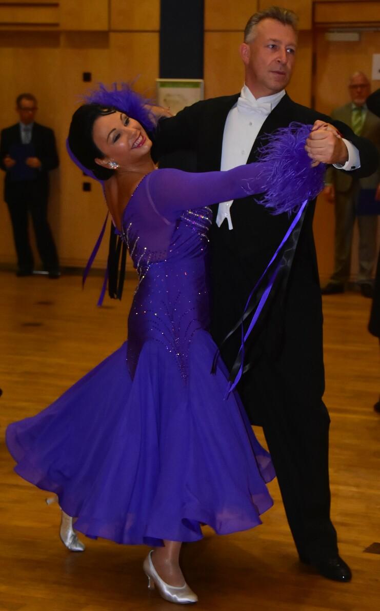 Tanzpaar in eleganter Kleidung tanzt in einem Saal; die Frau trägt ein lila Kleid mit Glitzer und Federn.