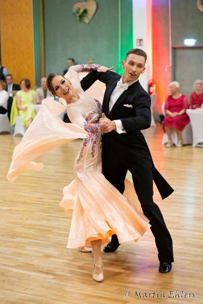 Paar tanzt in eleganter Kleidung auf einer Tanzfläche, umgeben von Zuschauern bei einem Tanzwettbewerb.