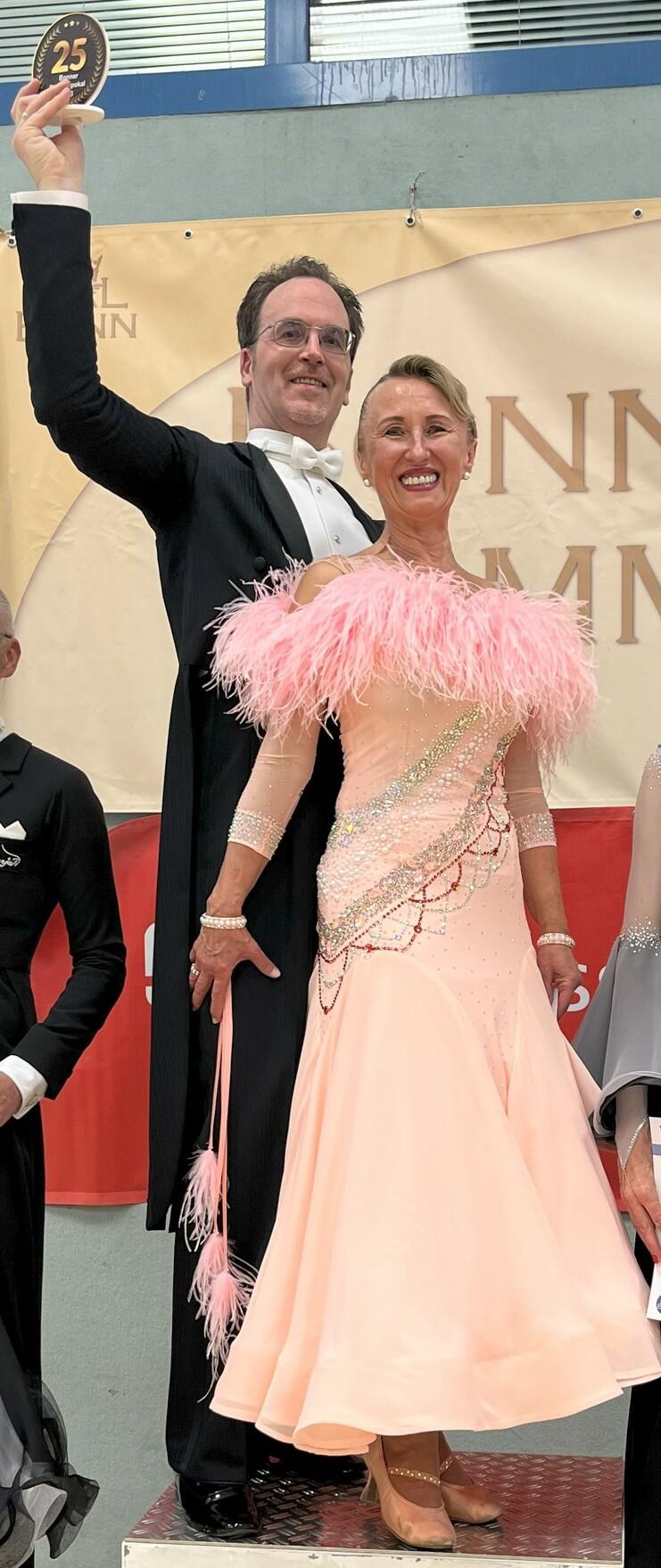 Tanzpaar auf dem Siegerpodest mit Urkunden, der Mann hält einen Pokal über dem Kopf. Sechs Tänzer in festlicher Kleidung.