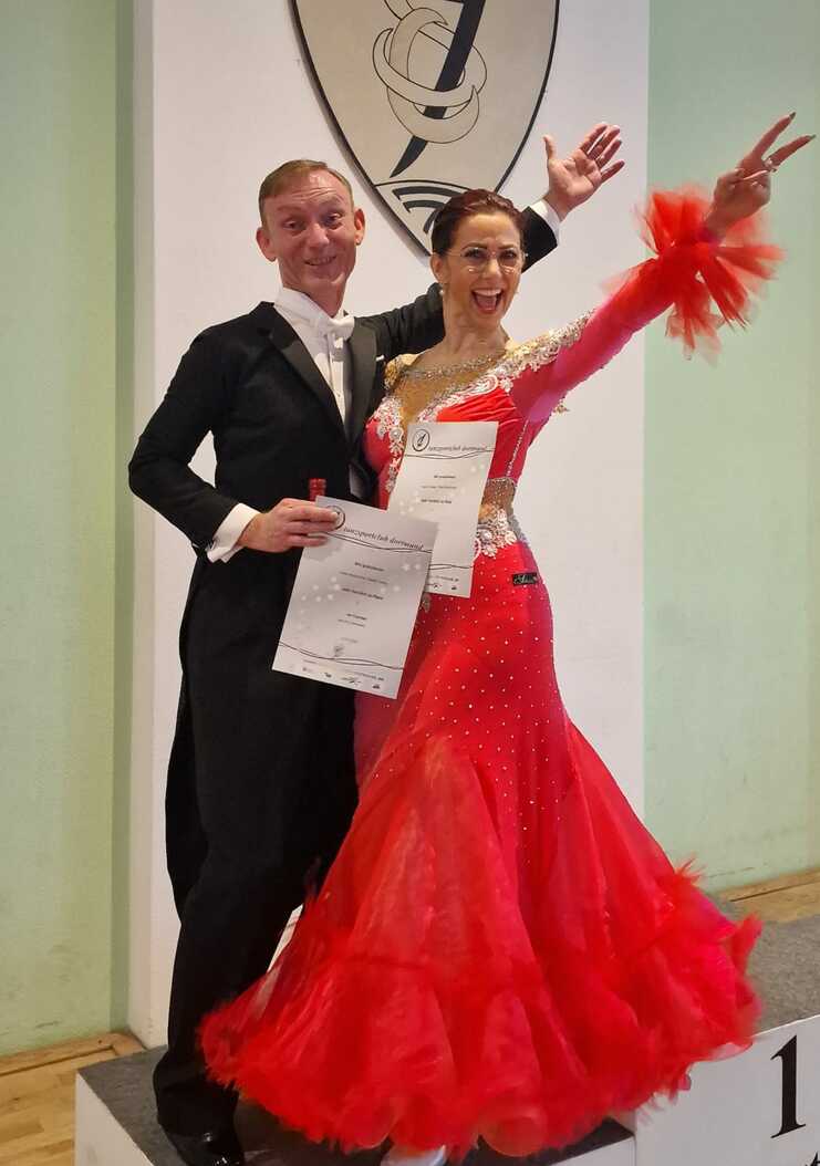 Tanzpaar in festlichen Kleidern steht auf dem Siegerpodest mit Urkunden, hinter ihnen das Logo des Tanzsportclubs Dortmund.
