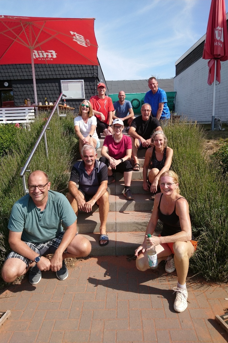 Gruppe von zehn Personen sitzt auf einer Treppe vor einem Gebäude mit Sonnenschirm und Grünpflanzen im Vordergrund.