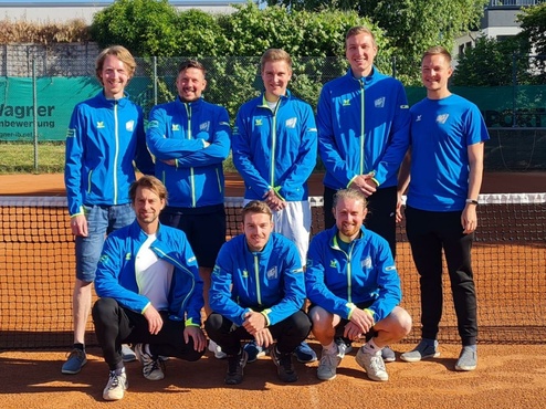 Gruppe von sieben TennisTrainern in blauen Jacken posiert vor einem Tennisplatz mit Netz und Hintergrundbewuchs.