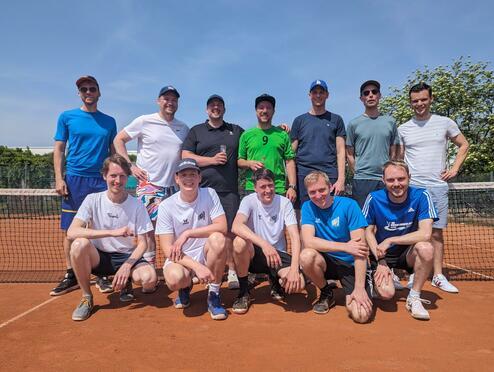 Gruppe von zehn Männern in Sportbekleidung auf einem Tennisplatz, lächelnd vor einem Netz im Freien.