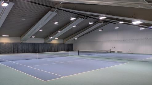 Innenansicht eines Tennisplatzes mit mehreren blauem Belag und Netz, unter einem hell erleuchteten schrägen Dach.