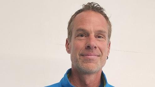 Mann in blauer Sportjacke mit Tennisschläger lächelt vor weißer Wand.