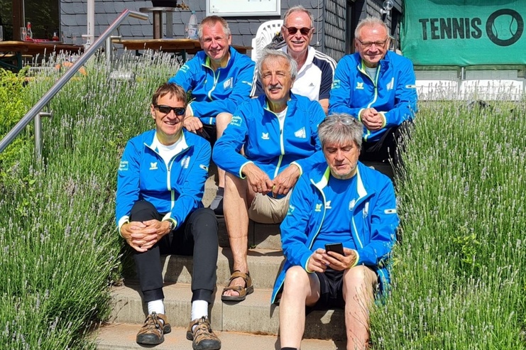 Sechs Personen in blauen Jacken sitzen auf einer Treppe, umgeben von Lavendel, vor einem Tennisplatz.