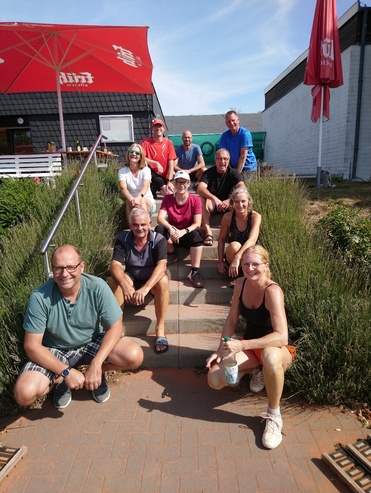 Gruppe von zehn Personen sitzt auf einer Treppe vor einem Geb&auml;ude mit Sonnenschirm und Gr&uuml;npflanzen im Vordergrund.
