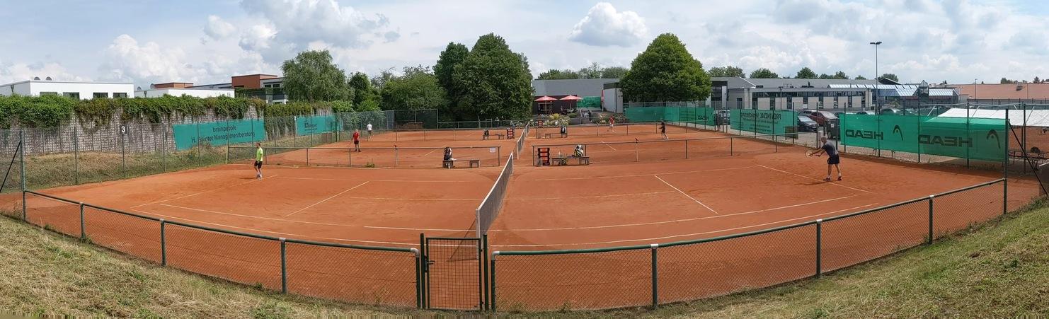 Panoramaaufnahme eines Tennisplatzes mit mehreren Spielern auf rotem Sand und umliegenden Gebäuden und Bäumen.